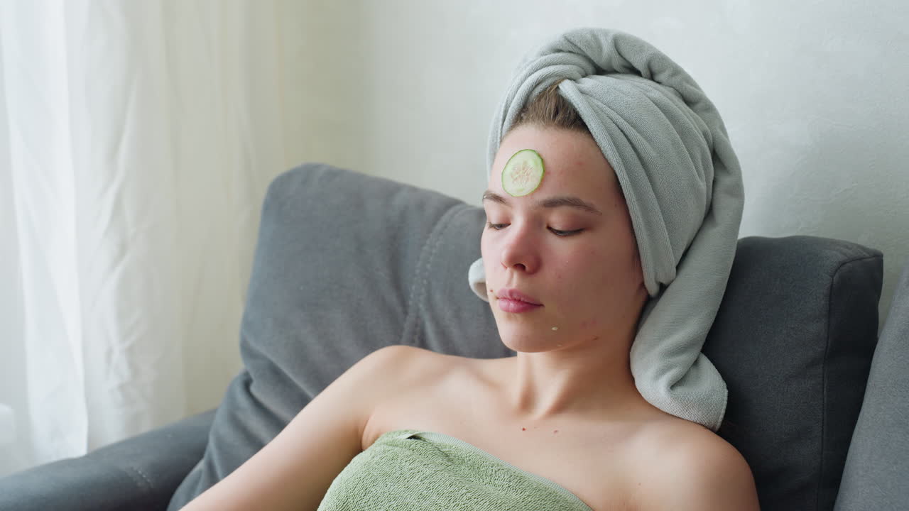 Relaxing woman wrapped in towel placed cucumber slice on forehead for skincare treatment, beauty ritual for refreshing and soothing skin, natural skincare therapy at home