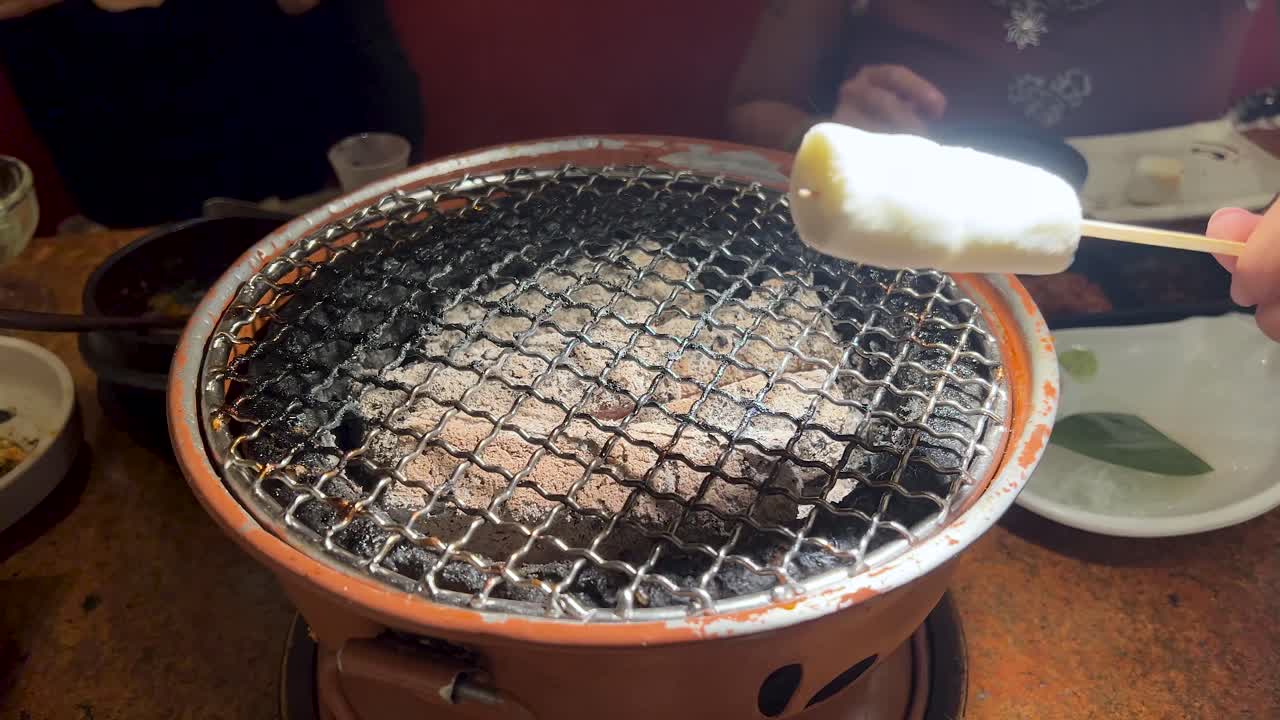 A hand rotates a marshmallow on a skewer above a small tabletop charcoal grill in a warmly lit restaurant, with diners and plates visible in the background