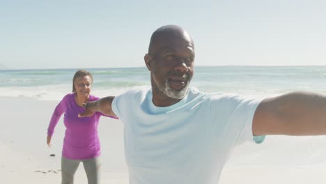 Smiling-senior-african-american-couple-practicing-yoga-on-sunny-beach