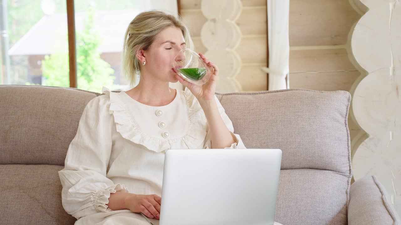 Woman relaxing on a sofa drinking green smoothie while working on a laptop.