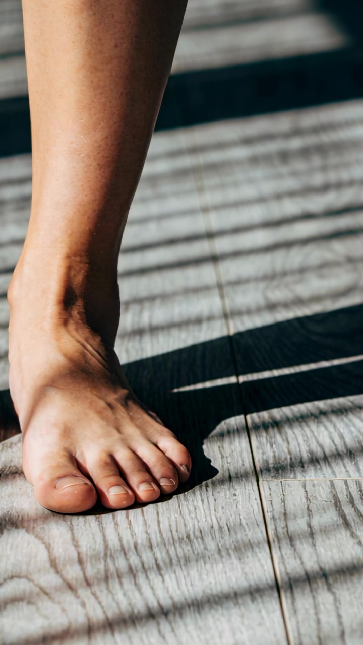 Vertical video: Lowering bare right foot contacting wood-plank floor to step, casting blind shadows