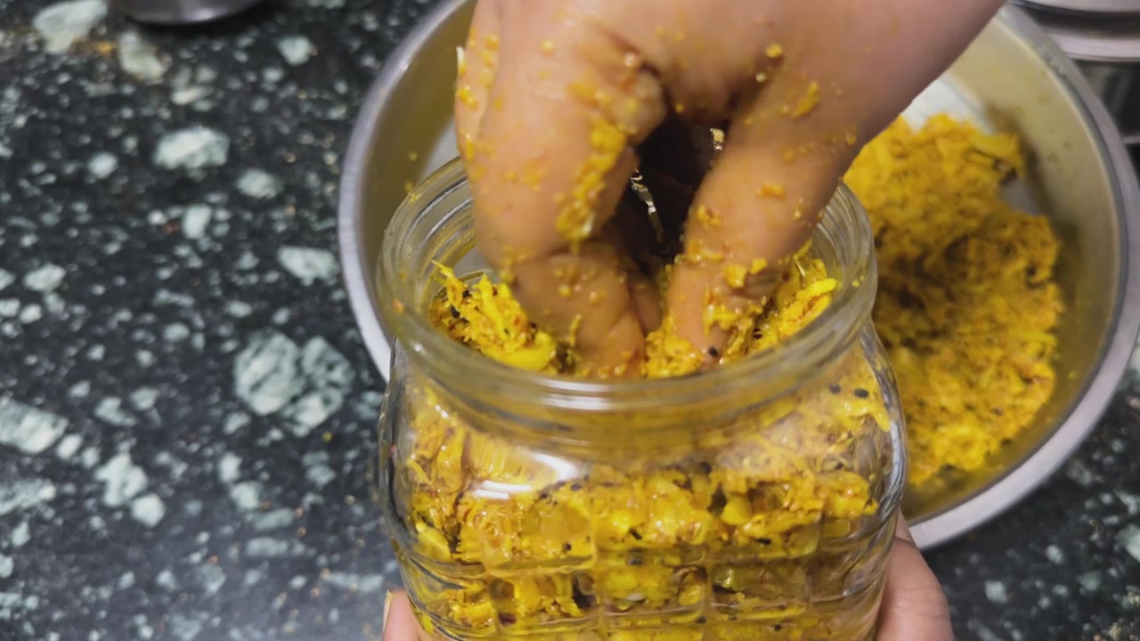 Detailed close view of hands filling freshly prepared shredded mango pickle with spices and mustard oil into a transparent glass jar during traditional Indian achar preparation