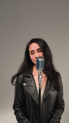 una hermosa vocalista con una chaqueta de cuero negra, sonriendo y cantando con los ojos cerrados, de pie en un micrófono vintage en un taburete alto. un guitarrista toca una guitarra roja y un tecladista trabaja un sampler rojo
