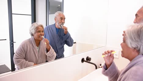 Happy-senior-biracial-couple-brushing-teeth-together-in-bathroom,-unaltered,-in-slow-motion