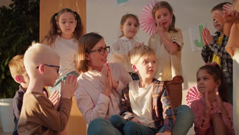 a female teacher with a bob hairstyle with glasses in a white shirt claps for a little blond boy along with the rest of the group of children in the pre-school club