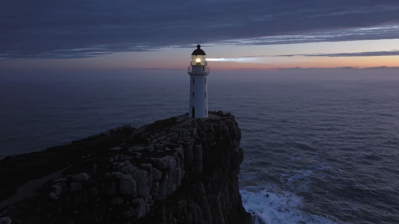 Aerial video of a lighthouse on a rocky cliff at dusk, capturing the serene ocean horizon