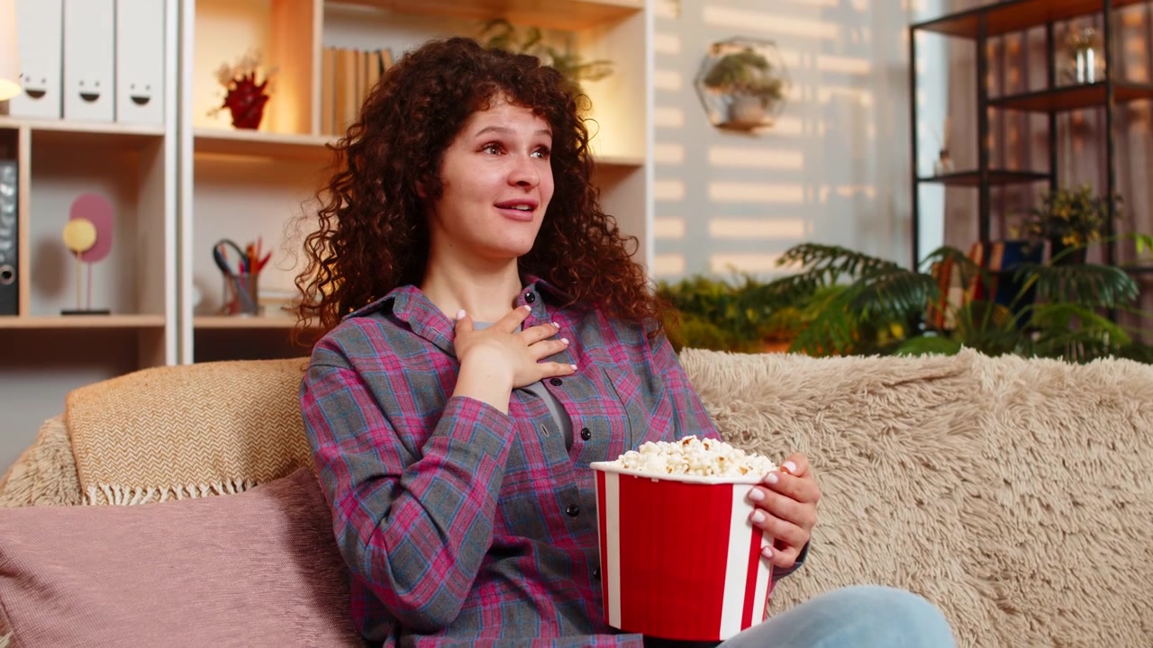 Ung kvinde på sofaen ser sjov film med popcorn og griner overrasket under hjemmebiograf-aften.