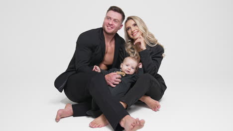 Happy couple in love sits on the floor with their baby beside. Caucasian parents with infant child in studio. White backdrop.