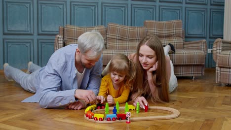 Family-of-mother,-father-with-daughter-child-girl-riding-toy-train-on-wooden-railway-at-home-room