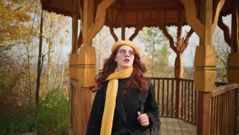 lady in black coat, yellow scarf, and beret stepping out of wooden gazebo in serene autumn park, adjusting bag strap, warm golden hues of trees complement rustic setting