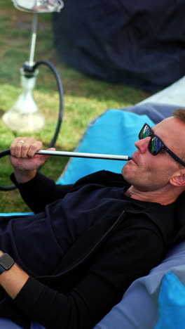 male hookah smoker lies in bean bag chair outdoors. man in sunglasses resting outside and looks at his laptop. view from above. vertical video