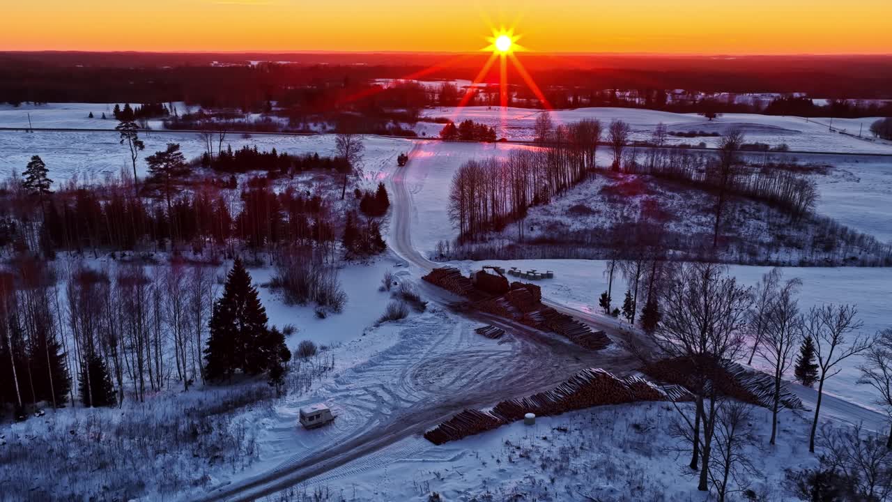 Drone kuvaa kultaista auringonnousun valoa, joka osuu puupinoihin lumisella Latvian maaseudulla, huurteen peittämine metsineen ja jäätyneine maaperineen talvella