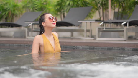 an attractive woman standing in chest-high water of a resort swimming pool looks around the hotel property