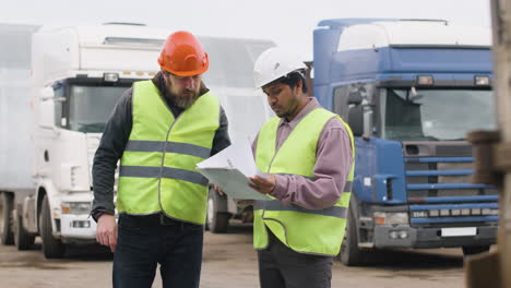 boss and worker wearing vests and safety helmets organizing a truck fleet in a logistics park while they consulting a document 2