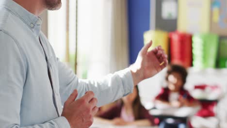 portrait of happy caucasian male teacher standing in classroom with children during lesson