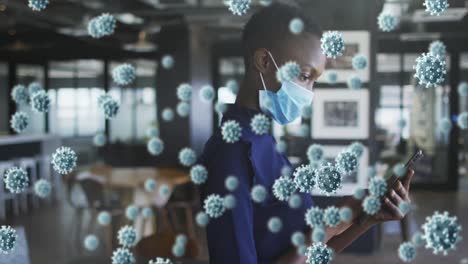 animation of covid 19 cells floating over woman in face mask using smartphone in office