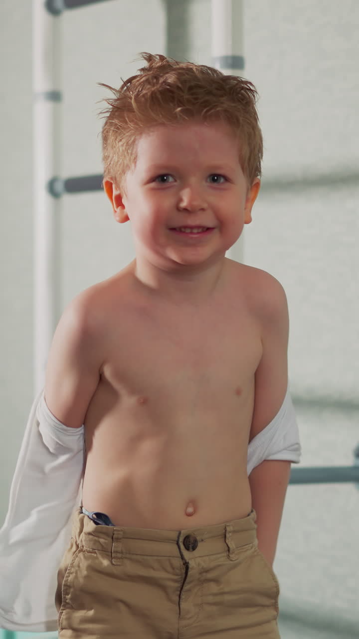 Smiling toddler boy takes off torn shirt standing with bare torso in empty domestic gym slow motion. Positive little child has fun at fighting training in class