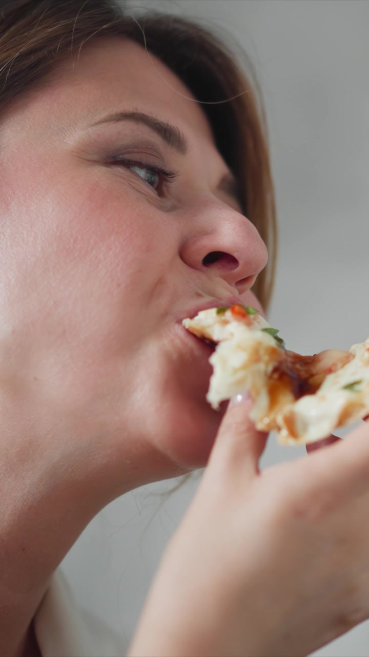 Close up of woman eating pizza. subject holds and takes bites from food with fine details like cheese stretchiness on display. Background is plain light gray emphasizing facial expressions
