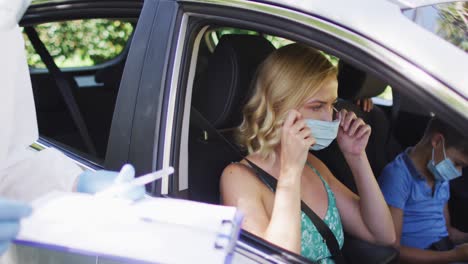 caucasian woman sitting in car, wearing face mask, having covid test done by medical worker outdoors