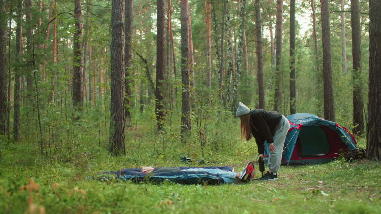 Man lays on tent bag resting as woman slightly bent pumps air into bag beside tent, surrounded by trees and soft daylight in peaceful forest camping scene