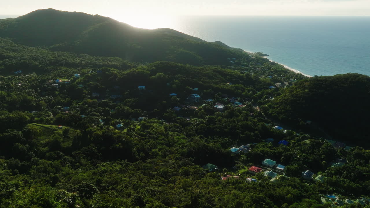 Drone moves forward above tropical hills at sunrise, revealing houses hidden in the forest and the sea in the background