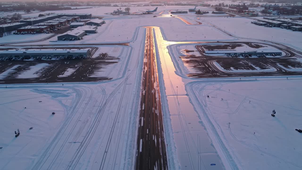 Aerial perspective captures snow-covered runway reflecting warm light, illustrating the evolving scene with airport structures and tranquil winter surroundings