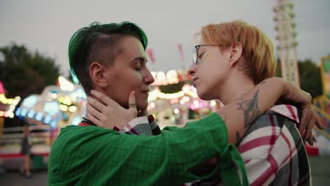 Close-up-shot-of-a-blonde-girl-with-short-hair-with-glasses-in-a-plaid-shirt-and-a-girl-with-short-green-hair-in-a-Green-shirt-hug-each-other-and-stroke-their-shoulders-during-their-date-in-the-amusement-park