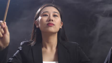 안개가 있는 검은 스튜디오에서 눈을 감고 몸짓을 하는 아시아계 지휘자 여성의 클로즈업