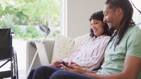 Happy-biracial-couple-in-living-room-using-laptop-and-smartphone,-her-crutch-and-wheelchair-by-couch