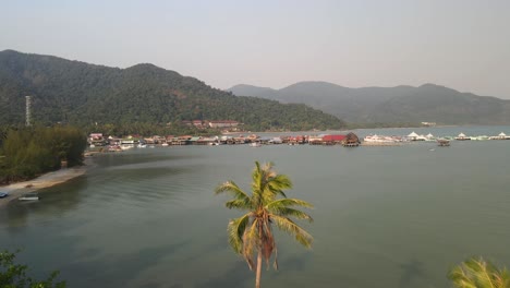 Disparo-Aéreo-Rápido-Sobre-Dos-Palmeras-A-Lo-Largo-De-La-Costa-De-Koh-Chang,-Tailandia-Con-El-Muelle-De-Pesca-Bang-Bao-En-La-Distancia