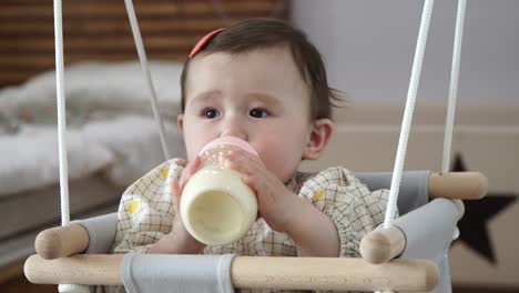 Baby-Girl-Drinking-Formula-Milk-By-Feeding-Bottle-While-Sitting-On-Wooden-Swing-Chair