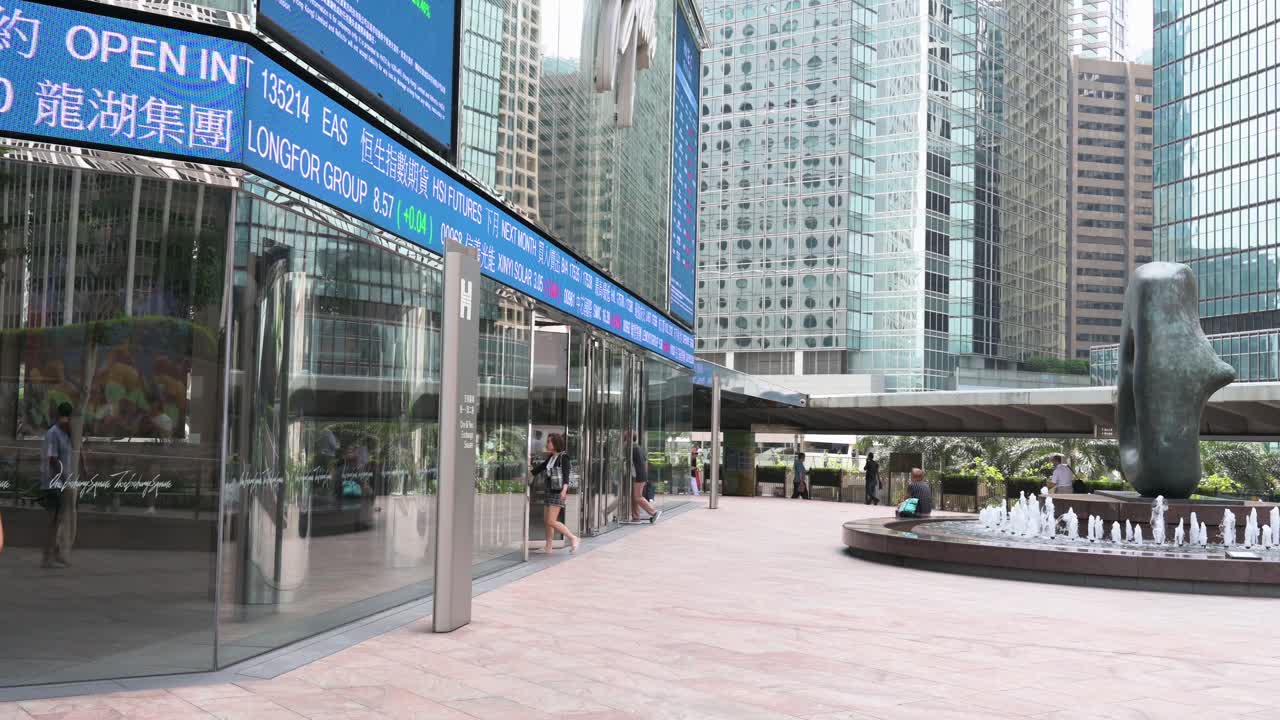 A low-angle view shows people moving past a dynamic screen at Exchange Square, home to the Hong Kong Stock Exchange (HKEX), which displays stock ticker symbols in Hong Kong's financial district.