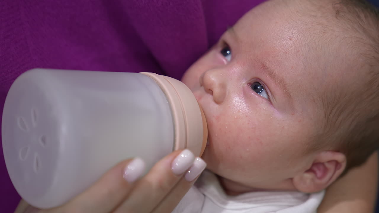 Beautiful sleepy baby suckling milk from a bottle. Mother's feeding her child holding him in hands. Close up.