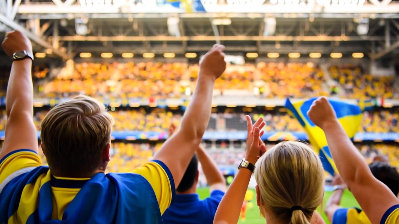 To jublende fans i lagtrøyer løfter armene, vifter med et lite svensk flagg på stadion og reagerer på banen.
