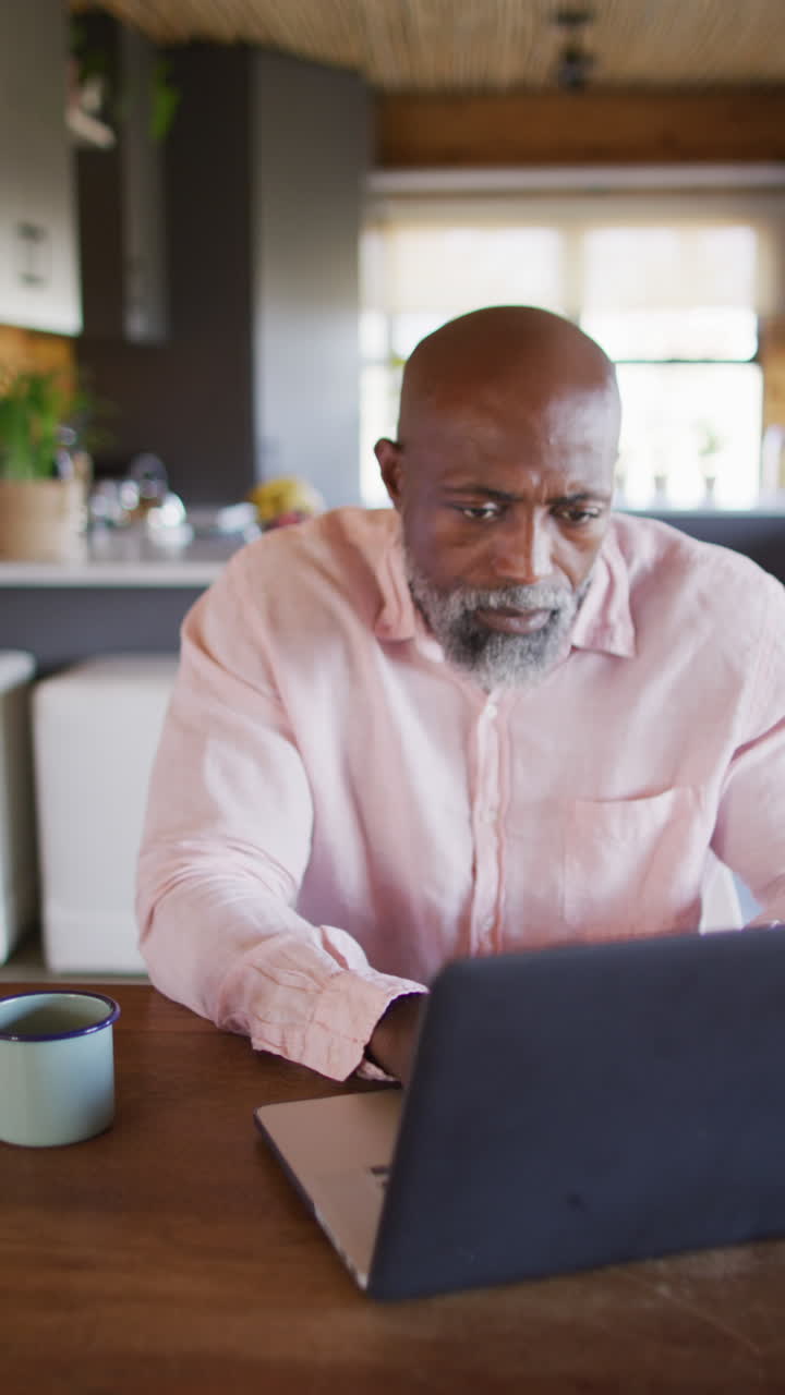 Vertical video of senior african american man spending time in log cabin and using laptop