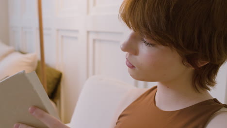 Vista-De-Cerca-De-Una-Chica-Sentada-En-Una-Silla-Leyendo-Un-Libro-En-El-Dormitorio