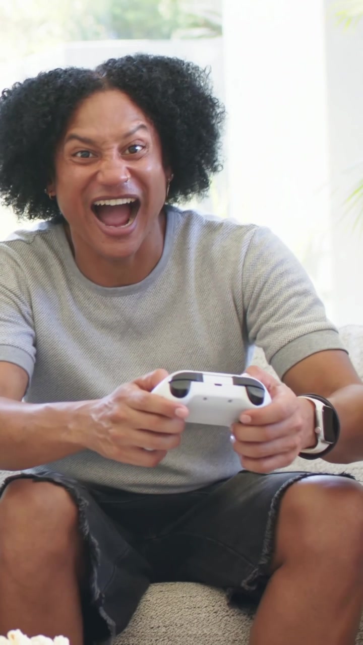 Vertical video: Cheering after game event, man gripping gamepad on couch in living room wearing tee