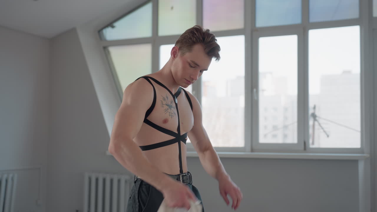 shirtless young student with black strap harness adjusts white shirt in bright modern room with large windows, natural daylight illuminating upper body as he focuses on adjusting clothing over torso