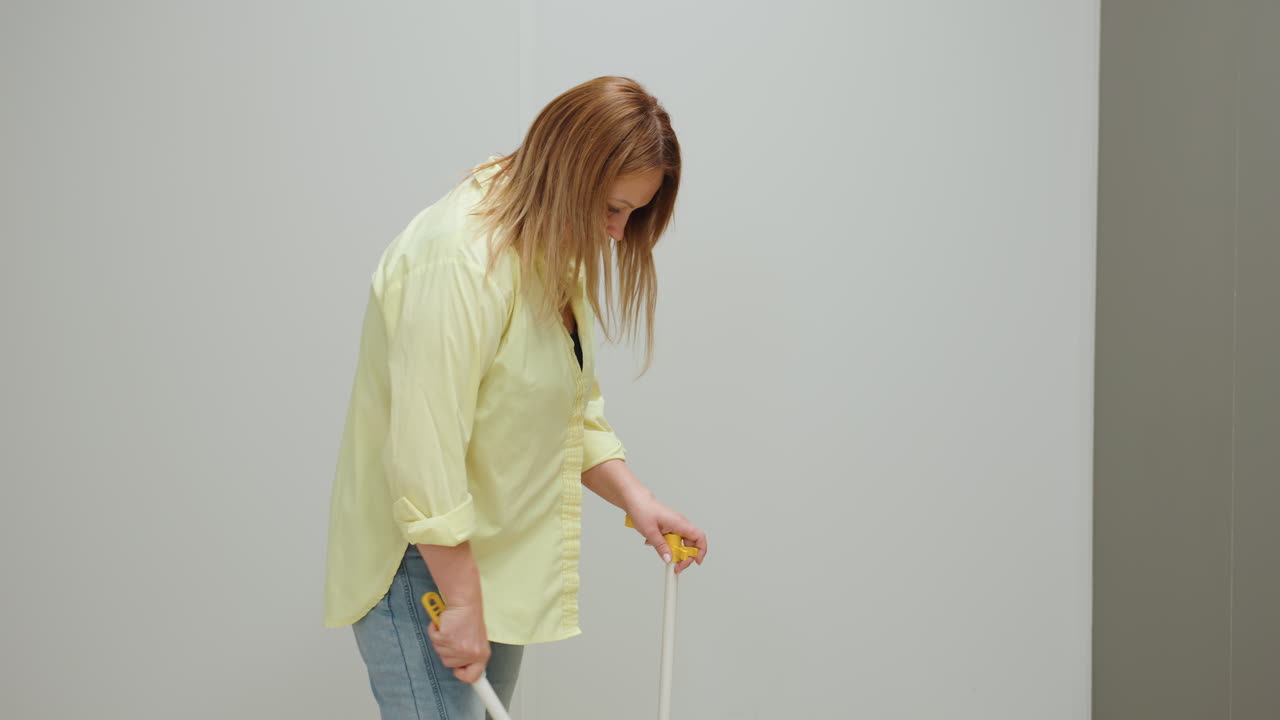 Medium shot shows laundry owner in yellow shirt, white sneakers and jeans sweeping tiled floor with broom and dustpan inside clean service room, maintaining hygiene, routine maintenance, tidy view