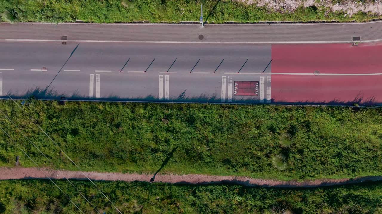 Ripresa dall'alto con drone che mostra una strada e un sentiero sterrato che corrono paralleli nell'inquadratura, con auto in movimento e una persona che cammina lungo il sentiero.