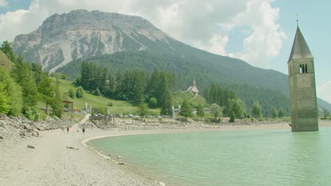 Ruhige-Aufnahme,-Menschen,-Die-An-Einem-Sonnigen-Tag-In-Italien-Am-Ufer-Des-Reschensees-Spazieren,-Im-Hintergrund-Der-Kirchturm-Von-Altgraun