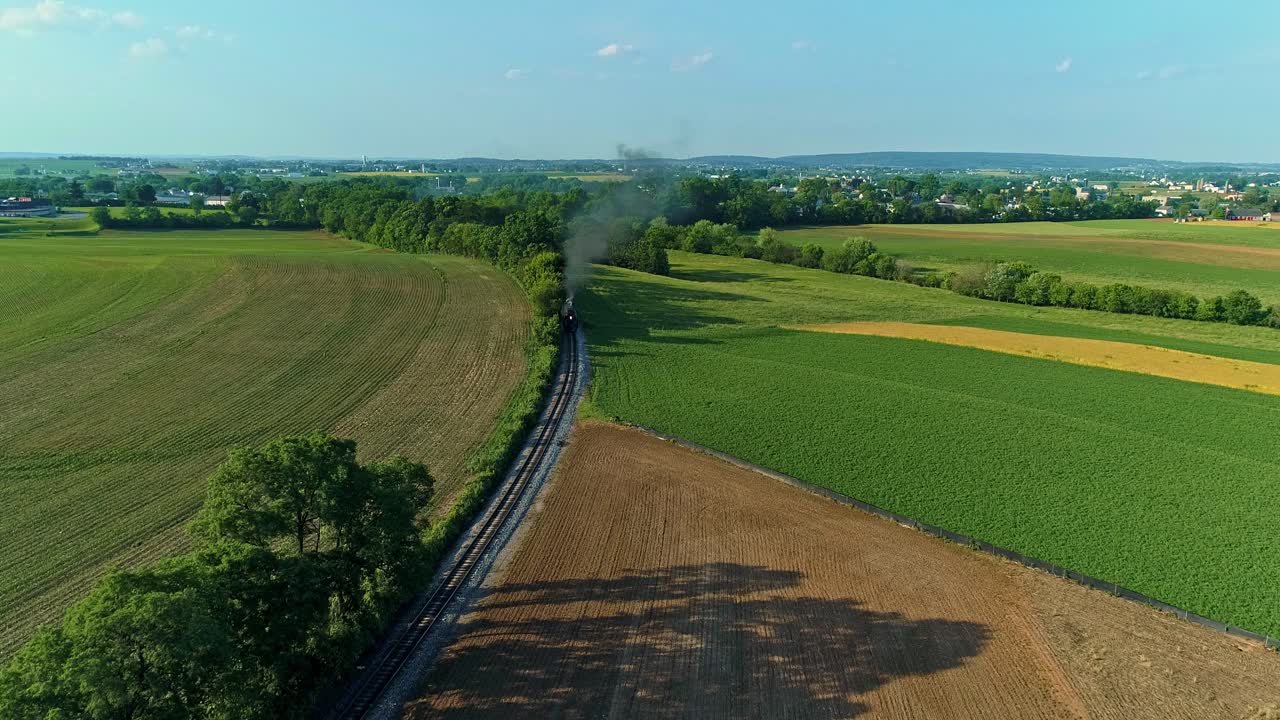 An Aerial View of a Steam Engine Puffing Smoke and Steam with Passenger Coaches Traveling on a Single Track Thru Trees and Farmland Countryside on a Beautiful Late Afternoon Golden Hour Spring Day