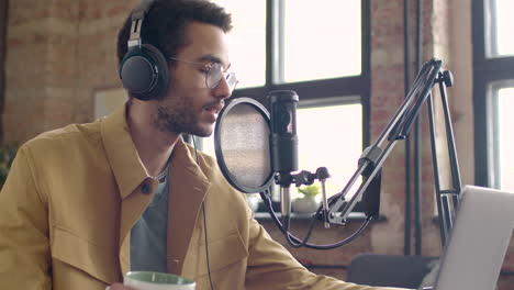 man recording a podcast wearing eyeglasses and headphones talking into a microphone while sitting on a table with laptop