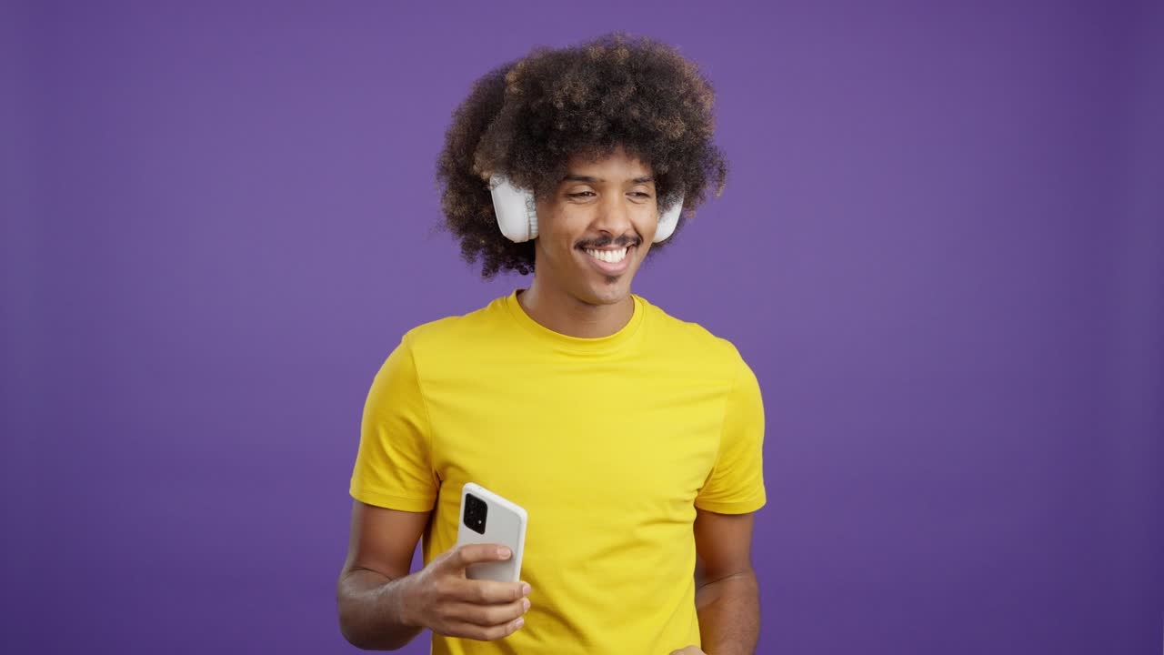 A man with afro hair and a mustache enjoys music with headphones and a smartphone on a purple background, smiling and dancing.