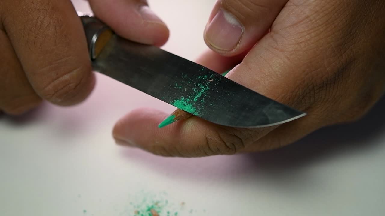 Footage of hands slowly sharpening a pencil and some coloured pencils with a sharp knife