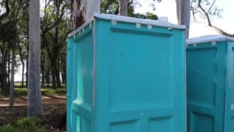 handheld-close-up-shot-of-aqua-green-blue-portable-restrooms-surrounded-by-trees-in-park