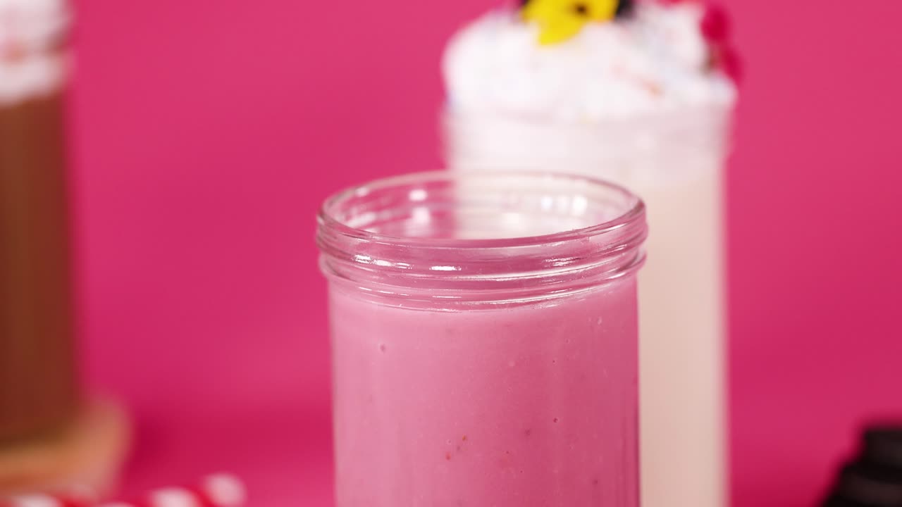 Pink milkshake in glass jar, whipped cream added, colorful dessert, bright pink background, studio lighting