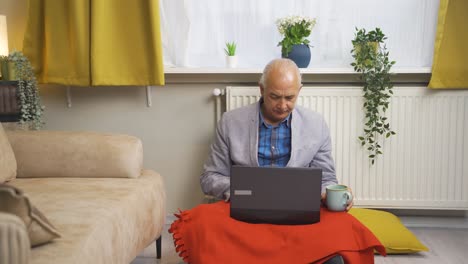 cold old man at home drinking coffee and using laptop.
