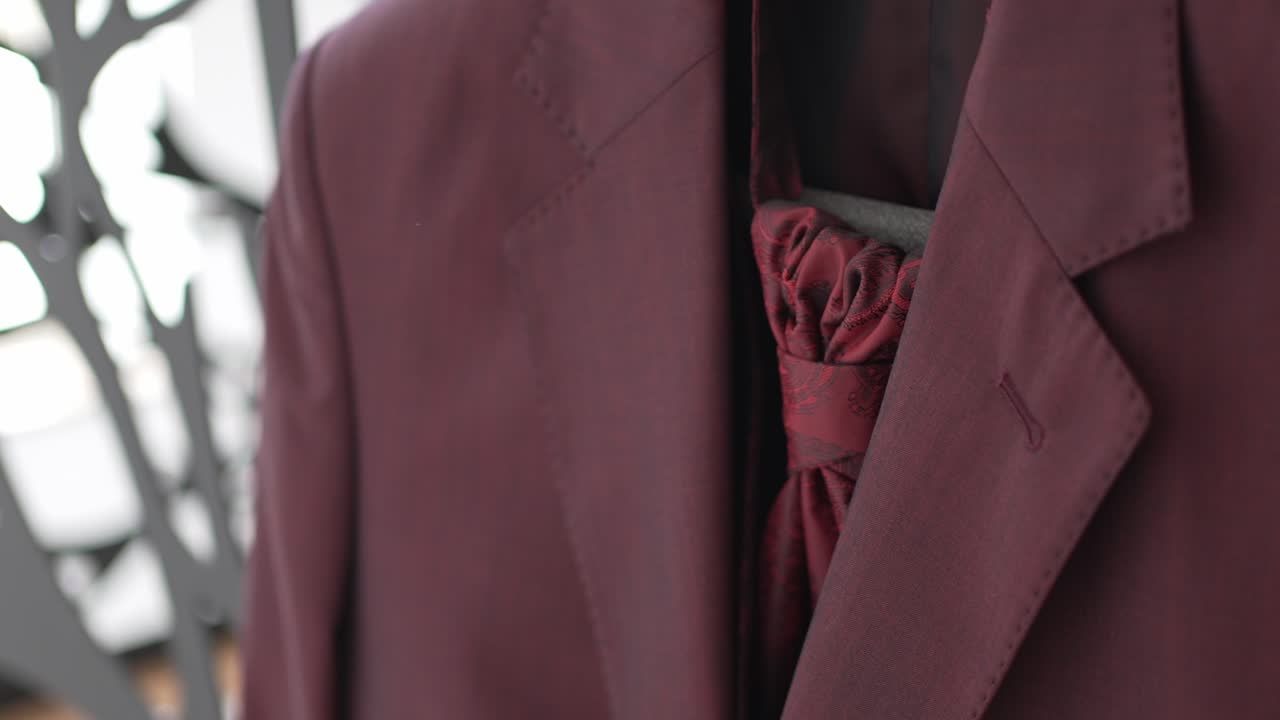 Close up of a burgundy groom’s suit with an elegant matching tie hanging indoors