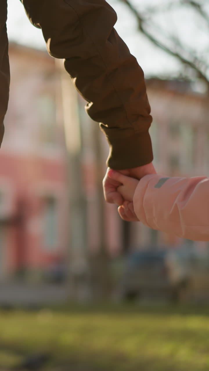 vista trasera de una niña en una gorra rosa y chaqueta, y un hombre en una chaqueta marrón, ambos con vaqueros azules, tomados de la mano mientras caminan por una calle soleada